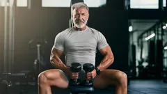 Starší muž ve fitku sedí na lavici s činkami, fitness a zdravý životní styl pro seniory.
