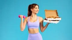 Žena ve sportovním oblečení drží činku a krabici pizzy na modrém pozadí