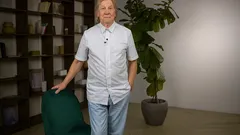 Starší muž stojí v místnosti, opírá se o židli, za ním knihovna a velká pokojová rostlina.