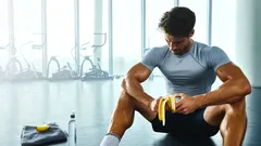 Muž ve fitku jí banán po cvičení, v pozadí fitness stroje, ručník a láhev vody.