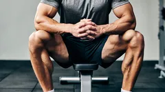 Muž sedící na lavici ve fitku, sportovní oblečení, svalnaté paže a nohy, fitness trénink